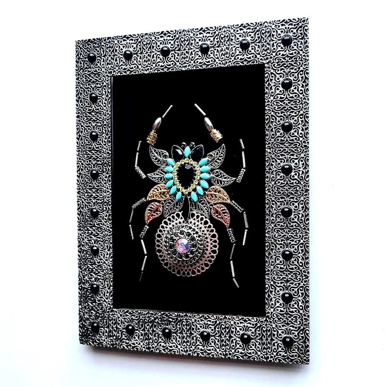 May include: Framed artwork of a spider, crafted from metal components. The spider features silver, gold, and copper elements, with turquoise accents and a central iridescent gem. The silver frame has a black pattern and is studded with black beads.
