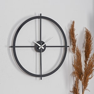 May include: A contemporary wall clock in matte black, featuring a circular frame with intersecting lines and silver hands. The clock has a black central dial. The diameter of the clock is approximately 61 cm.