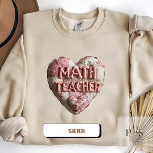 May include: Sand-colored sweatshirt featuring a heart-shaped design with the words "MATH TEACHER" in pink, with a smaller heart below. The design has a quilted appearance. The word "SAND" is displayed in a white rectangle at the bottom.