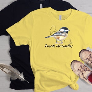 May include: A yellow t-shirt with a black outline of the state of Maine and a black outline of the state of Massachusetts. A black and white illustration of a bird is centered on the shirt with the text "Poecile atricapillus" below the bird.