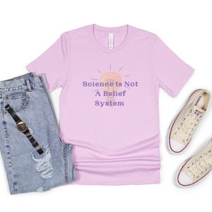 May include: A light pink t-shirt with a lightbulb graphic and the text "Science is Not A Belief System". The shirt is paired with blue jeans and white sneakers.