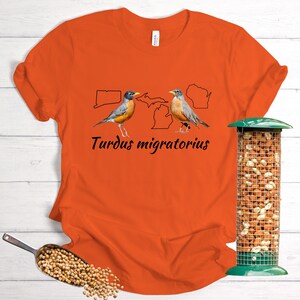 May include: Orange t-shirt with a black outline of the United States, Michigan, and Connecticut. Two robins are perched on the states with the text "Turdus migratorius" below.