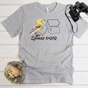 May include: A gray t-shirt with a yellow bird illustration and the text "Spinus tristis" on the front. The bird is perched on a branch with two outlines of the states of Washington and Montana behind it.