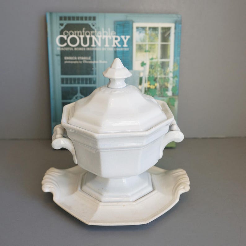 May include: A white, lidded soup tureen with a matching base. The tureen has a geometric design and handles. The base has a scalloped edge. A book titled "Comfortable Country" is in the background.
