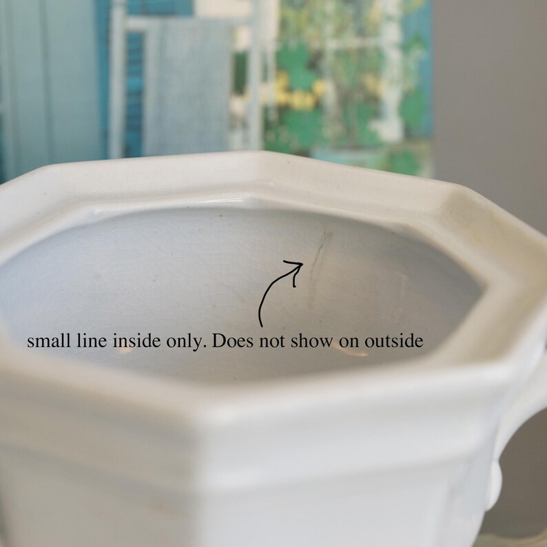 May include: Close-up of a white ceramic pitcher with an octagonal rim. The interior has a small line, as indicated by an arrow and text. The background is blurred, showing hints of blue and green.