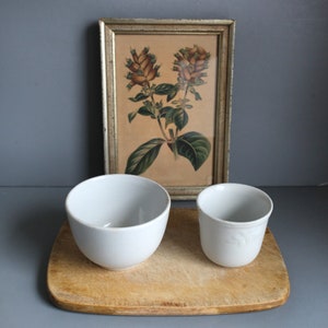 May include: Two white ceramic bowls, one larger than the other, are sitting on a wooden cutting board. The bowls are empty. Behind the bowls is a framed print of a botanical illustration of a flower with brown and green leaves.