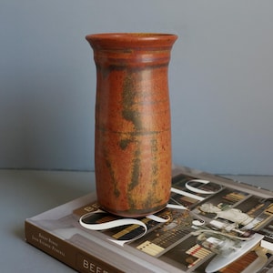 May include: A cylindrical ceramic vase with a flared rim, featuring a mottled glaze in shades of burnt orange, rust, and deep brown with subtle vertical streaking and a smooth, slightly tapered body design.