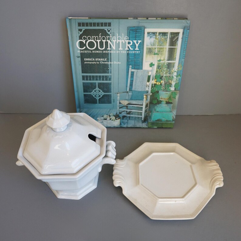 May include: A white ceramic soup tureen with a lid and a matching serving platter. The tureen has a geometric design and handles. A book titled "Comfortable Country" is in the background.