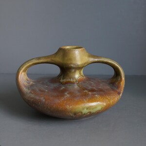 May include: A low-profile ceramic vase featuring a wide, rounded body and two prominent, arched handles extending from the central neck. The surface displays a mottled glaze with blended tones of earthy brown, burnt orange, and muted olive green.
