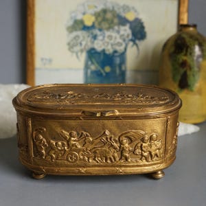 May include: A gold-colored, oval-shaped jewelry box with a detailed relief design. The box features a hinged lid and small feet. The relief depicts figures, chariots, and landscapes. The box is made of metal and has a vintage aesthetic.