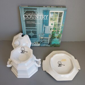 May include: White ceramic serving dish with lid and handles, alongside a matching plate. The dish has a hexagonal shape and a black and white emblem. A book titled "Comfortable Country" is in the background.