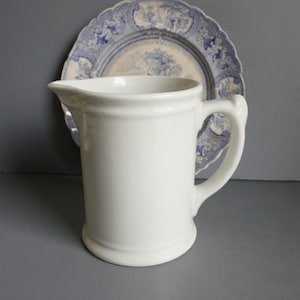 May include: A white ceramic pitcher with a curved handle and a pouring spout. Behind the pitcher is a blue and white decorative plate with a scalloped edge and a landscape design. The pitcher is a classic design.