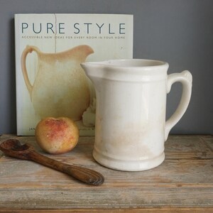 May include: A cream-colored ceramic pitcher with a smooth finish and a side handle, positioned next to a round peach and a dark brown wooden spoon. Behind these items stands a book titled PURE STYLE with a picture of a pitcher on the cover.