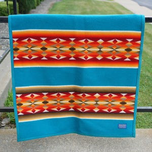 May include: A turquoise wool blanket featuring horizontal bands of geometric patterns in shades of red, orange, yellow, and brown. The design includes repeating diamond shapes and thin stripes, with a small rectangular label in the bottom corner.