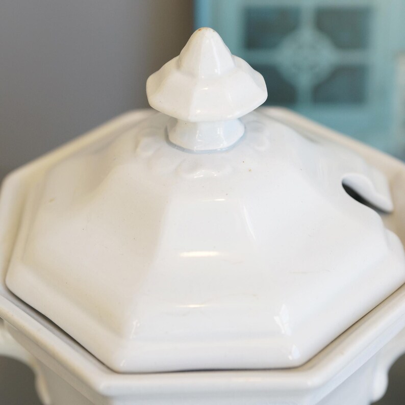 May include: Close-up of a white ceramic soup tureen lid. The lid is octagonal with a small, decorative finial on top. A small opening is visible for a spoon. The tureen appears to be vintage or antique.