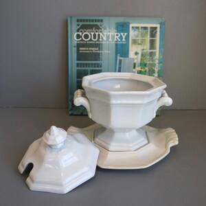 May include: White ceramic soup tureen with lid and serving dish. The tureen has two handles and a pedestal base. The lid has a small knob and a slot for a spoon. The serving dish has a scalloped edge. A book titled "Comfortable Country" is in the background.