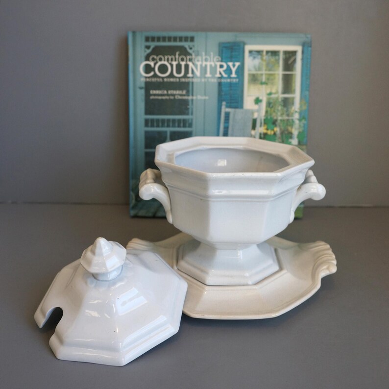 May include: White ceramic soup tureen with lid and serving dish. The tureen has two handles and a pedestal base. The lid has a small knob and a slot for a spoon. The serving dish has a scalloped edge. A book titled "Comfortable Country" is in the background.