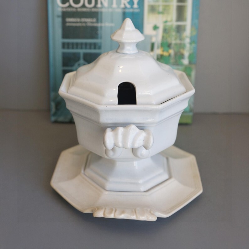 May include: White ceramic soup tureen with a lid and a matching octagonal base. The tureen has a handle on each side and a small opening in the lid for a ladle. The base has a decorative edge.