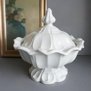 May include: A white ceramic lidded serving dish with a pedestal base. The dish has a scalloped edge and a decorative finial on the lid. The design is classic and elegant, suitable for serving food.