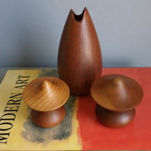 May include: A tall, teardrop-shaped wooden vase with a pointed rim, accompanied by two small, mushroom-shaped wooden containers with conical lids. The items are crafted from a medium-brown wood with visible grain patterns and a smooth finish.