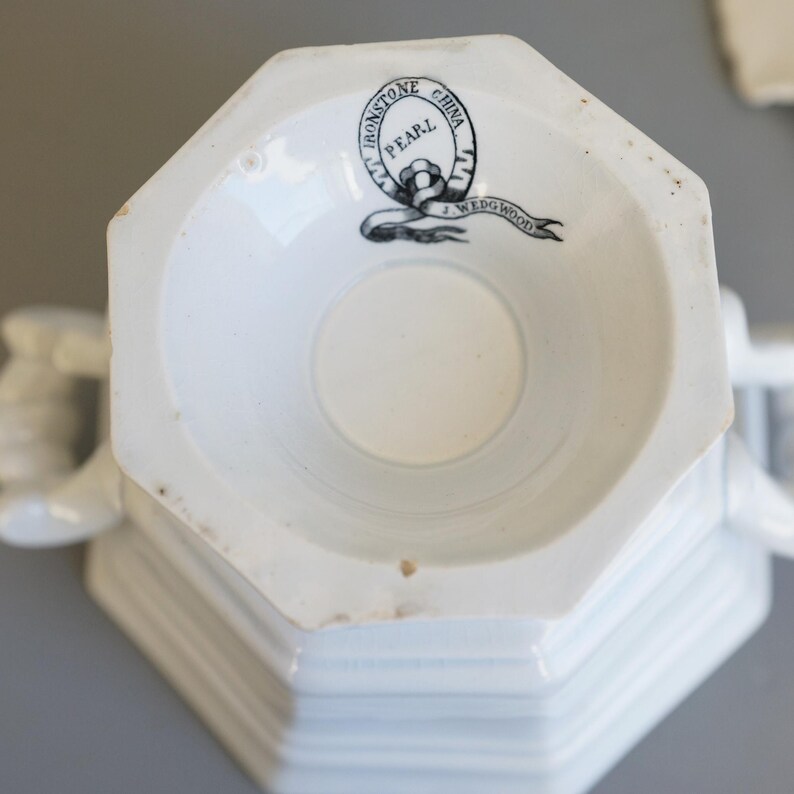 May include: A white, octagonal teacup base with a circular indentation. The bottom of the cup features a black and white logo that reads "Ironstone China" and "Wedgwood". The cup has a handle on each side.