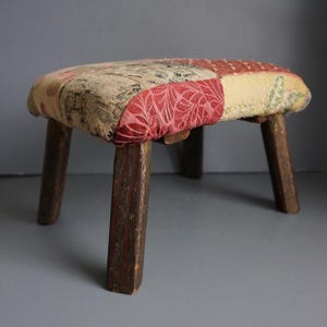 May include: A small, handcrafted stool with a patchwork fabric seat in shades of red, beige, and green. The seat rests on four sturdy, dark brown wooden legs. The stool's design suggests a vintage or antique aesthetic.
