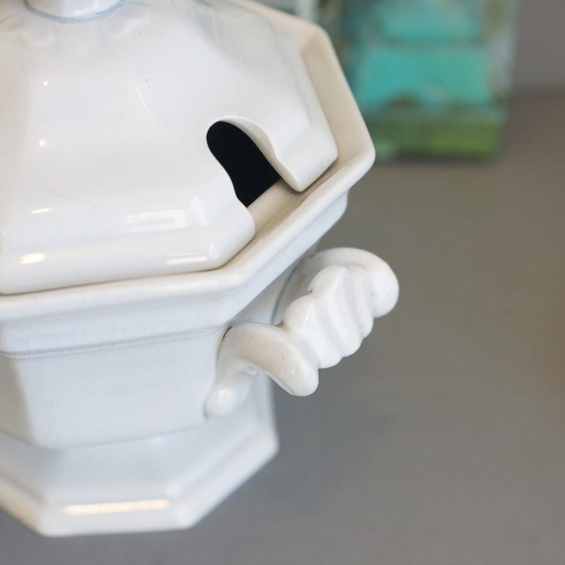 May include: White ceramic soup tureen with a lid and handle. The tureen has an octagonal shape and a small opening in the lid for a ladle. The handle is designed to look like a hand.