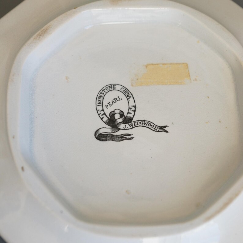 May include: Close-up of the bottom of a white ceramic dish. The dish has an octagonal shape. The center features a black and white logo that reads "Ironstone Pearl China J. Wedgwood."