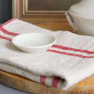 May include: A small, white ceramic dish sits on a folded, cream-colored linen towel with two red stripes. The dish is round with a slightly raised rim. The towel rests on a wooden surface, with a blurred background.