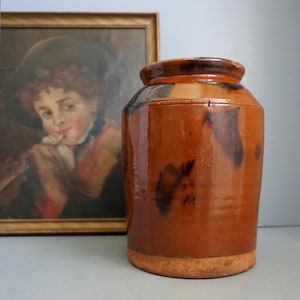 May include: A cylindrical stoneware crock featuring a glossy, mottled brown glaze with darker brown patches. The vessel has a thick, rounded rim and a wide opening, with an unglazed, light tan base that shows the natural texture of the fired clay.