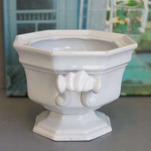 May include: A white ceramic planter with an octagonal shape and a pedestal base. The planter features decorative handles and a smooth, glossy finish. The design is classic and elegant, suitable for displaying small plants or flowers.