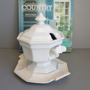 May include: A white ceramic soup tureen with a lid and a matching plate. The tureen has a geometric design and a handle. The lid has a small opening. The background includes a book with the title "comfortable COUNTRY".