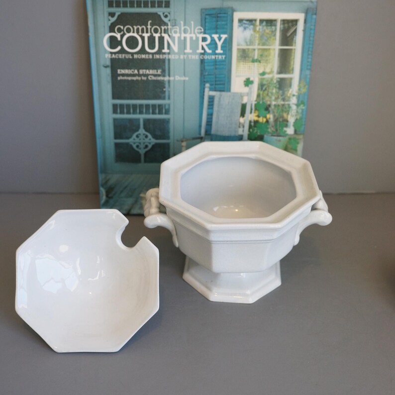 May include: White ceramic serving bowl with lid, featuring an octagonal shape and two side handles. The lid has a notch for a ladle. The bowl is set against a backdrop of a magazine with the title "Comfortable Country".