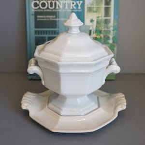 May include: White ceramic lidded soup tureen with a matching serving platter. The tureen has a geometric design with handles and a decorative finial on the lid. The platter has a scalloped edge.