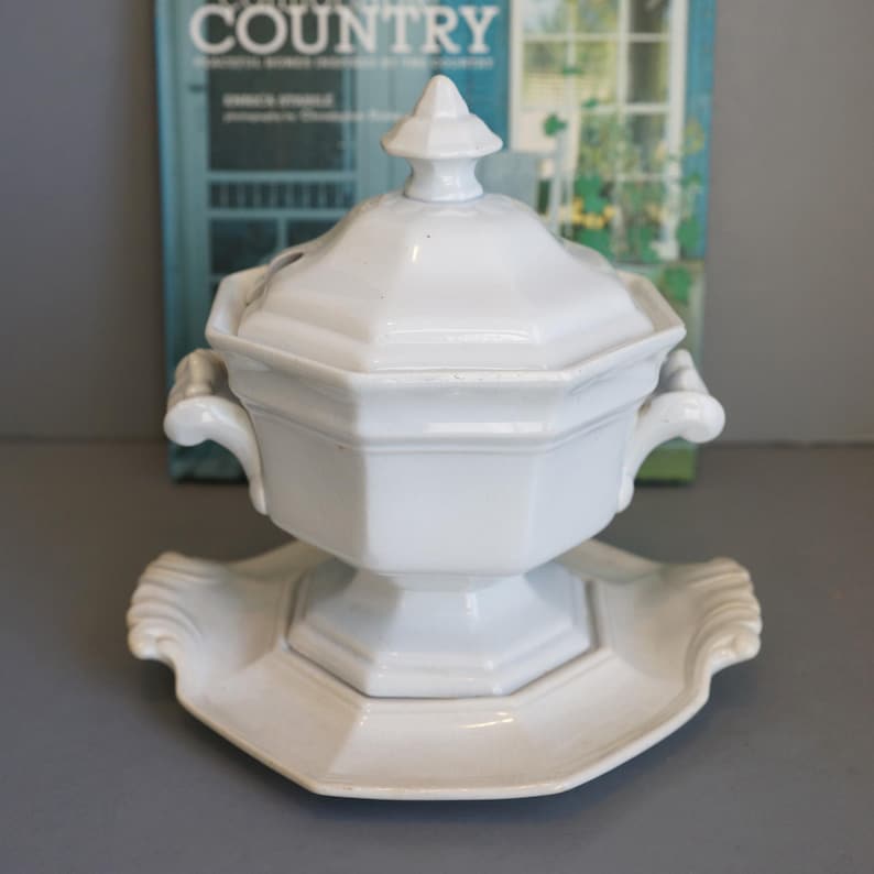 May include: White ceramic lidded soup tureen with a matching serving platter. The tureen has a geometric design with handles and a decorative finial on the lid. The platter has a scalloped edge.