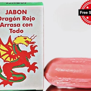 May include: A bar of pink soap next to a white box with green trim. The box features the words "JABON Dragón Rojo Arrasa con Todo" and a red dragon illustration. The soap is oval-shaped and has a smooth surface.