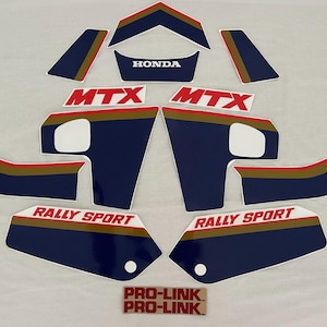 Könnte beinhalten: Ein Satz Motorradaufkleber in Blau, Gold und Rot mit den Worten "MTX", "RALLY SPORT", "HONDA" und "PRO-LINK" darauf gedruckt.