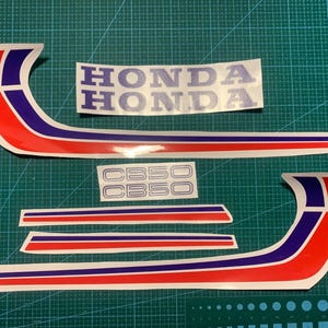 Könnte beinhalten: Satz Honda Motorradaufkleber in Rot, Weiß und Blau. Enthält das Honda-Logo und CB50-Markierungen. Die Aufkleber sind auf einer grünen Schneidematte angeordnet.