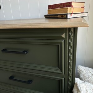 May include: A dark green dresser with two drawers and black drawer pulls. The dresser has a decorative trim around the edges. The top of the dresser is a light wood color.