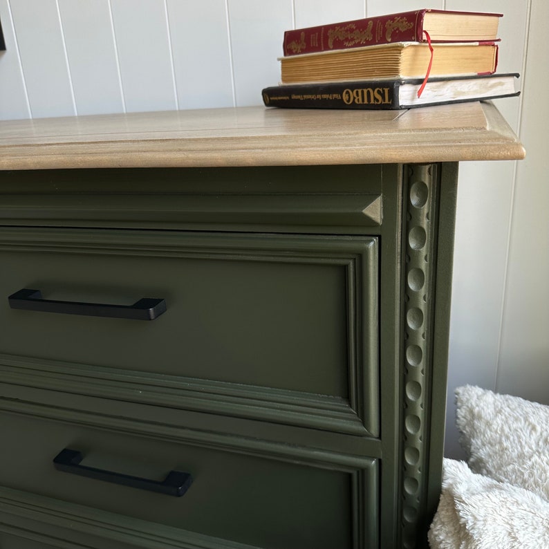 May include: A dark green dresser with two drawers and black drawer pulls. The dresser has a decorative trim around the edges. The top of the dresser is a light wood color.