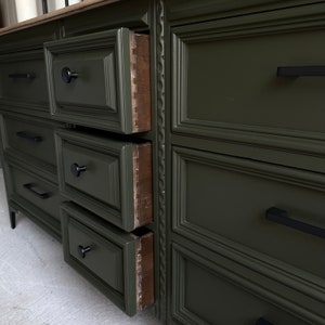 May include: A dark green dresser with six drawers, each with a black metal handle. The top drawer is open, revealing the wood interior.
