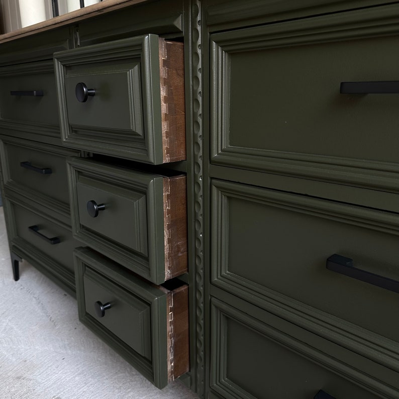 May include: A dark green dresser with six drawers, each with a black metal handle. The top drawer is open, revealing the wood interior.