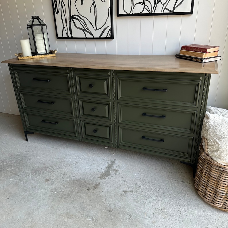 May include: A dark green dresser with eight drawers and black hardware. The dresser has a wood top and sits on black legs. The dresser is in front of a white wall with two black and white framed pictures above it.