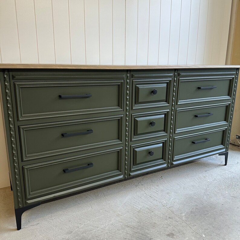 May include: A dark green dresser with eight drawers and black hardware. The dresser has a wood top and black legs.