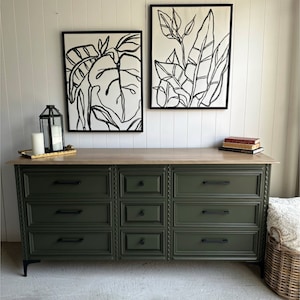 May include: A dark green dresser with six drawers and black hardware. The dresser has a wood top and sits on black legs. Two black and white framed prints of leaves hang on the wall behind the dresser.