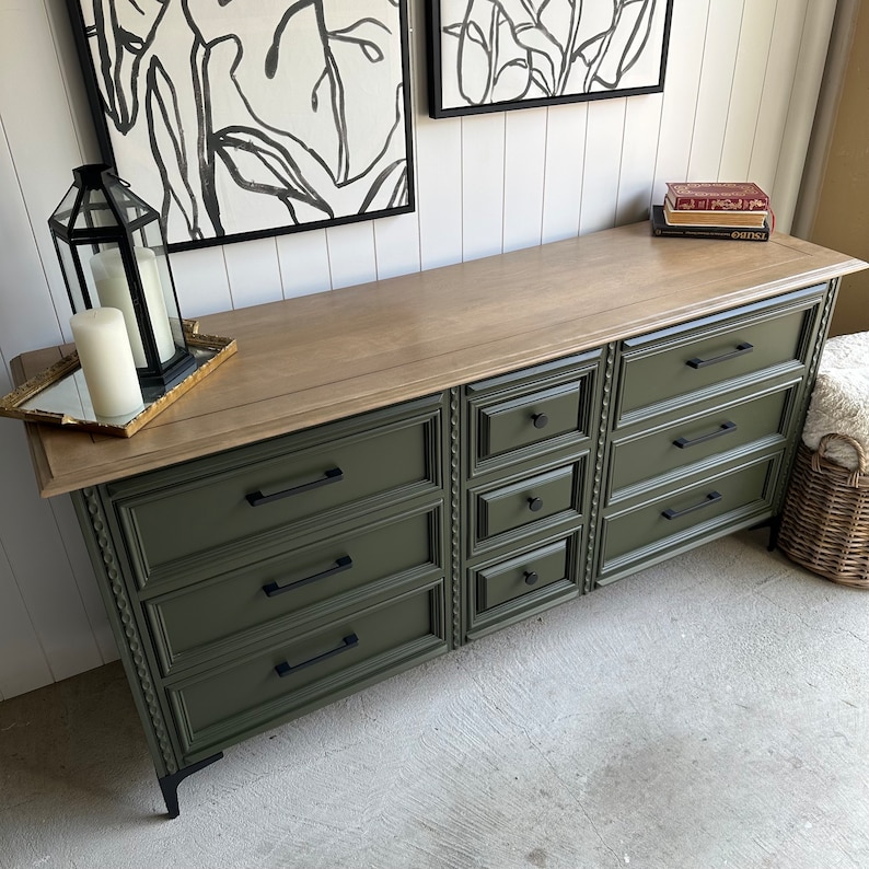 May include: A dark green dresser with eight drawers and black hardware. The dresser has a light wood top and sits on black legs. The dresser is against a white wall with two black and white framed pictures above it.