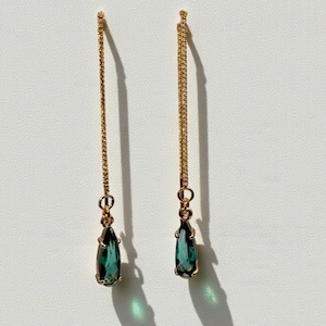 May include: A pair of gold-colored drop earrings. Each earring features a delicate gold chain suspending a teardrop-shaped, teal-colored gemstone. The earrings are displayed against a white background, with shadows cast by the sunlight.