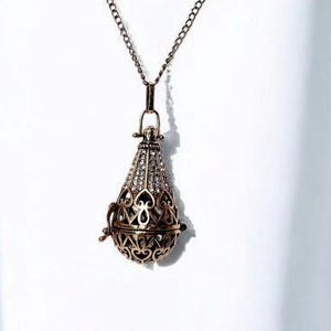 Puede incluir: Un delicado collar de color dorado con un colgante en forma de lágrima. El colgante está finamente diseñado con un patrón de filigrana y tiene una pequeña abertura en la parte superior.