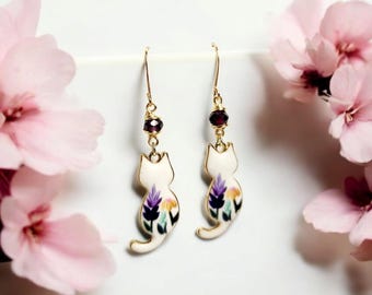 Puede incluir: Un par de pendientes colgantes de oro con dijes de gato de porcelana blanca con diseños florales y detalles en morado y amarillo. Cada dije tiene una pequeña cuenta de piedra preciosa morada encima.