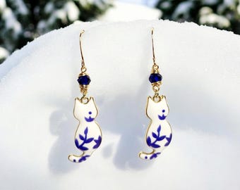 Puede incluir: Un par de pendientes colgantes con un gato de porcelana blanca con diseños florales azules y un acento de cristal azul oscuro.